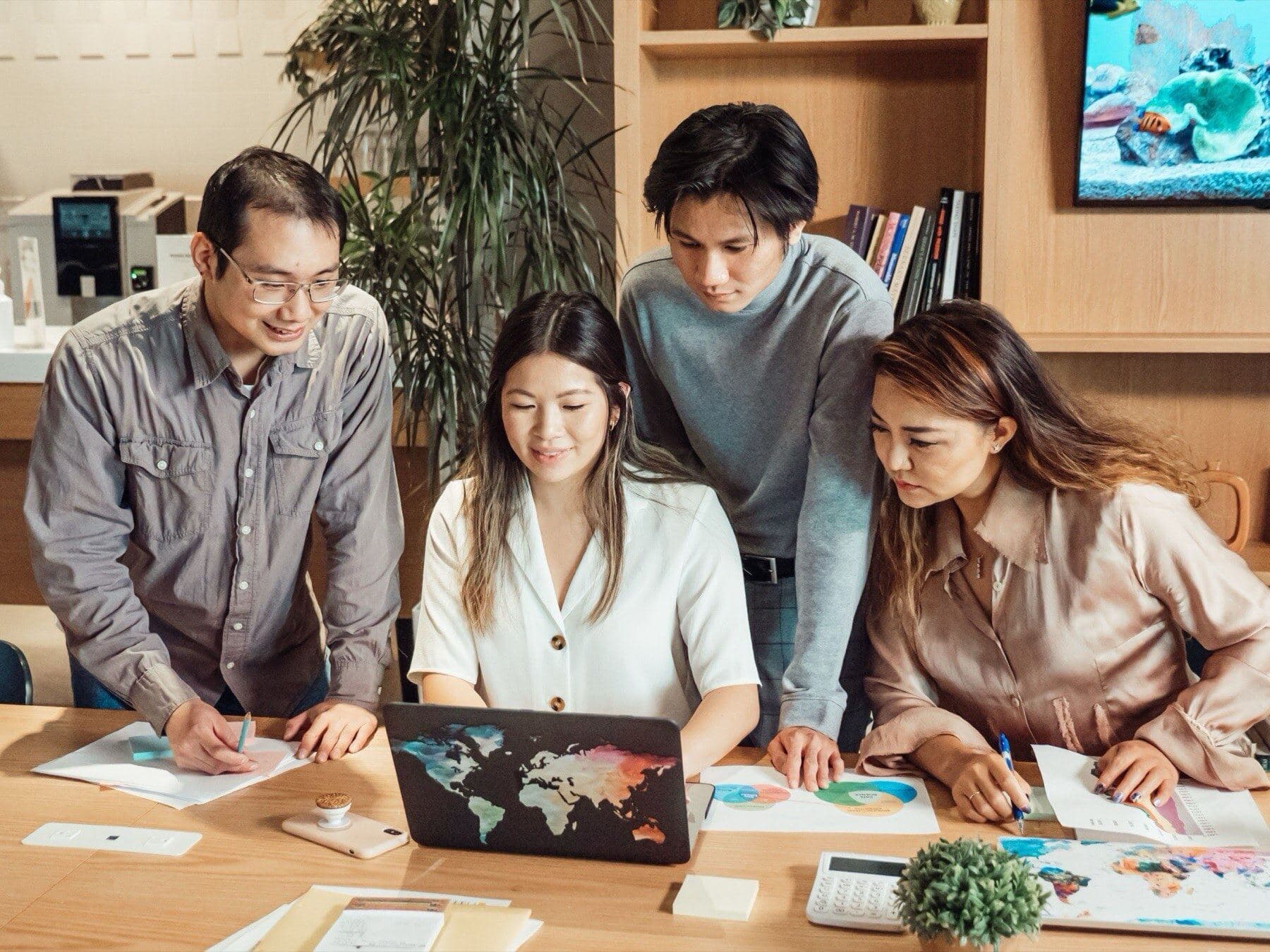 Four colleagues gather around a laptop printed with a world map, reviewing charts and notes spread across the table.