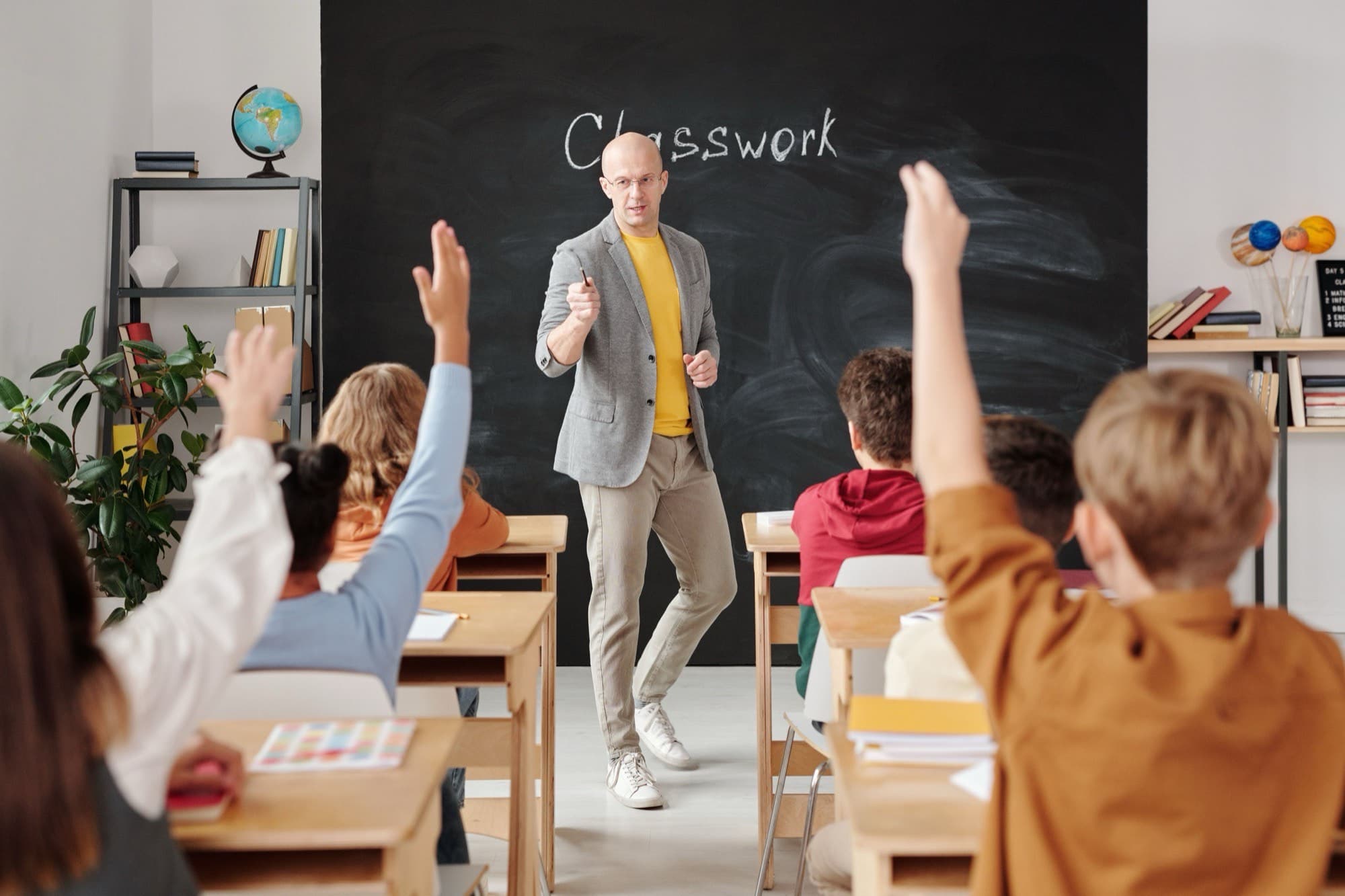 Children raise their hands as an educator walks toward them from the chalkboard, in a light, plant-filled classroom.