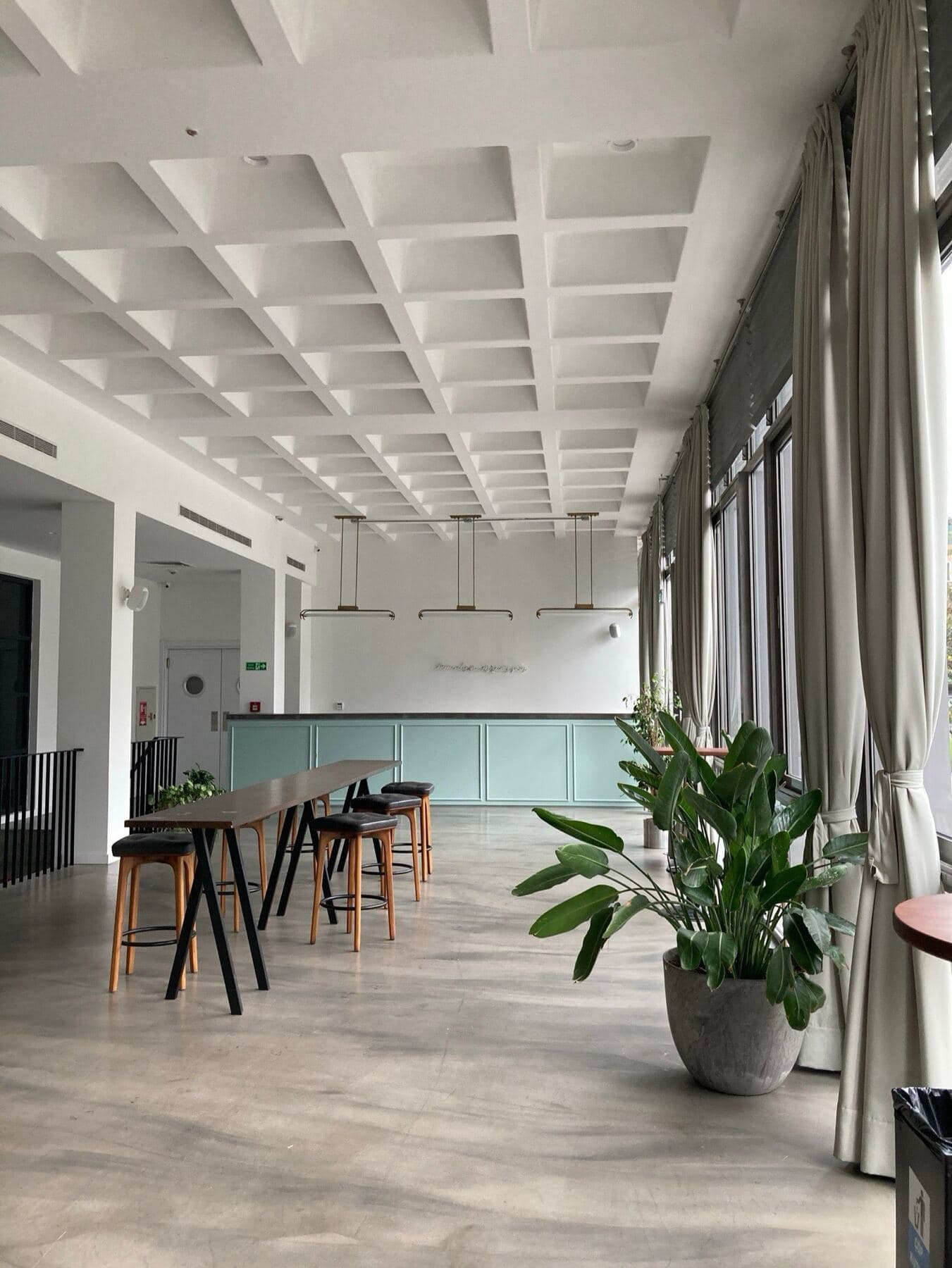 A quiet, sunlit learning venue with a long high table and stools beside floor-length windows.