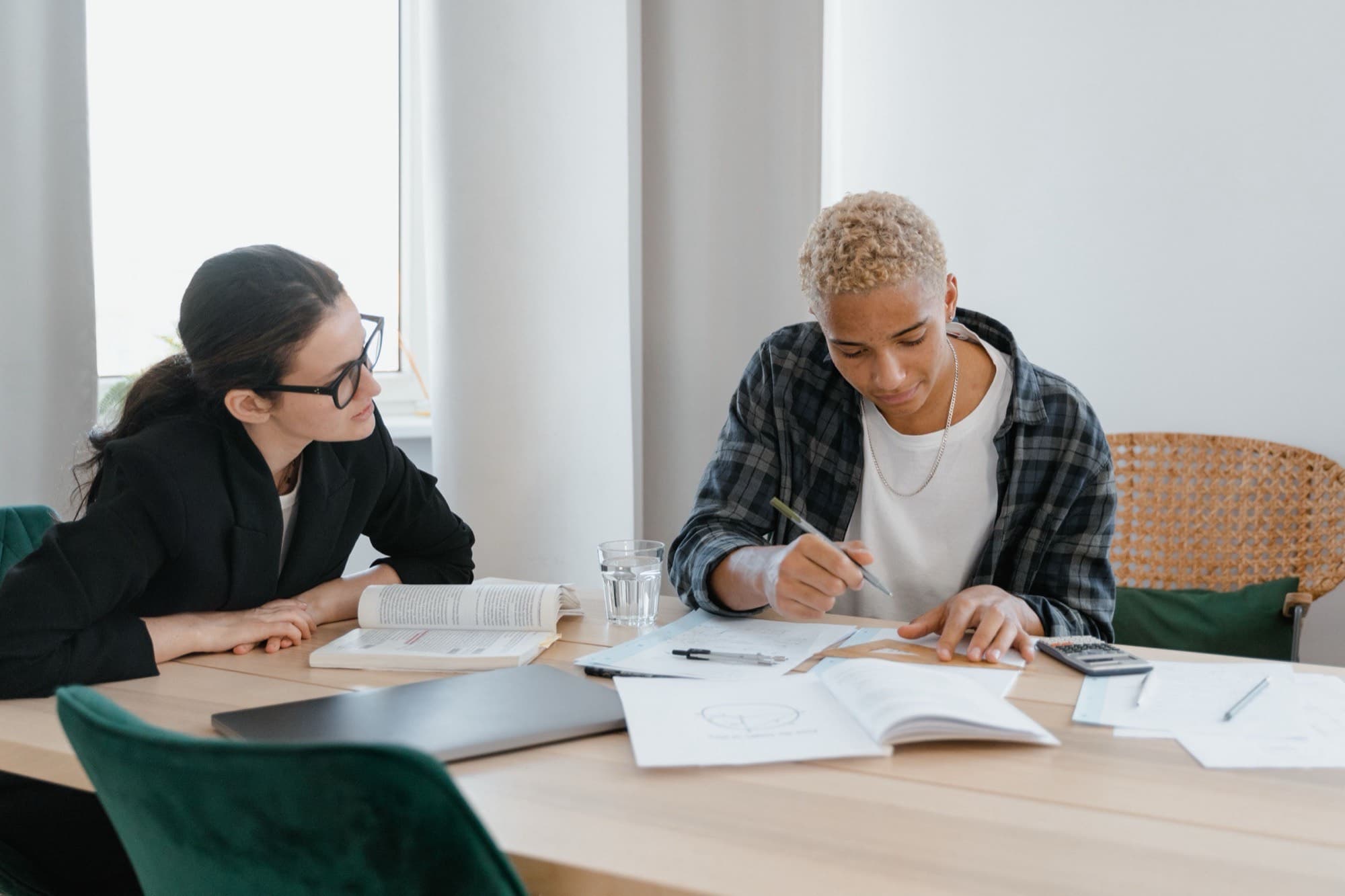 An educator in glasses sits beside a young man at a bright wooden table, watching closely as he works through a geometry problem on paper.