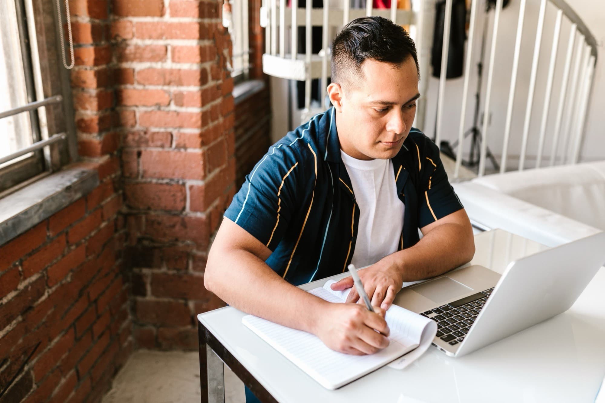 A man takes handwritten notes beside an open laptop at a white desk in a brick-walled loft.