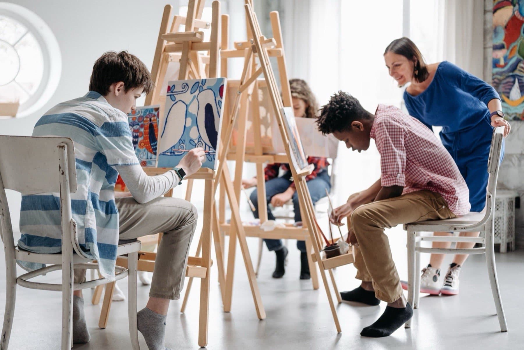 Young painters work at wooden easels in a local community studio while an educator leans in to look at one student's canvas.