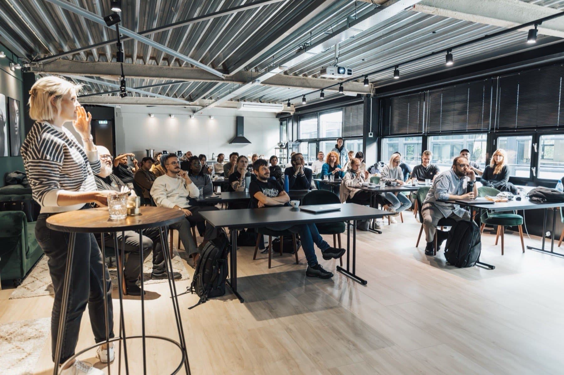 A facilitator leads a training session from a high table at the front of a loft room while a team of professionals listens from long tables.