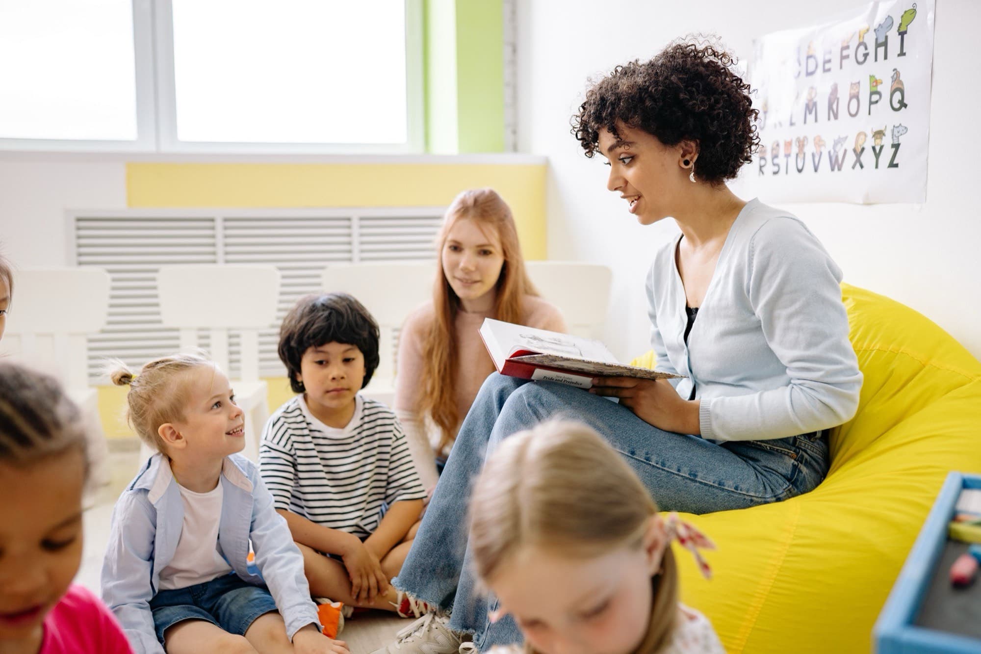 An educator on a yellow beanbag reads aloud to young children sitting on the floor, one girl smiling up at her beneath an alphabet chart.