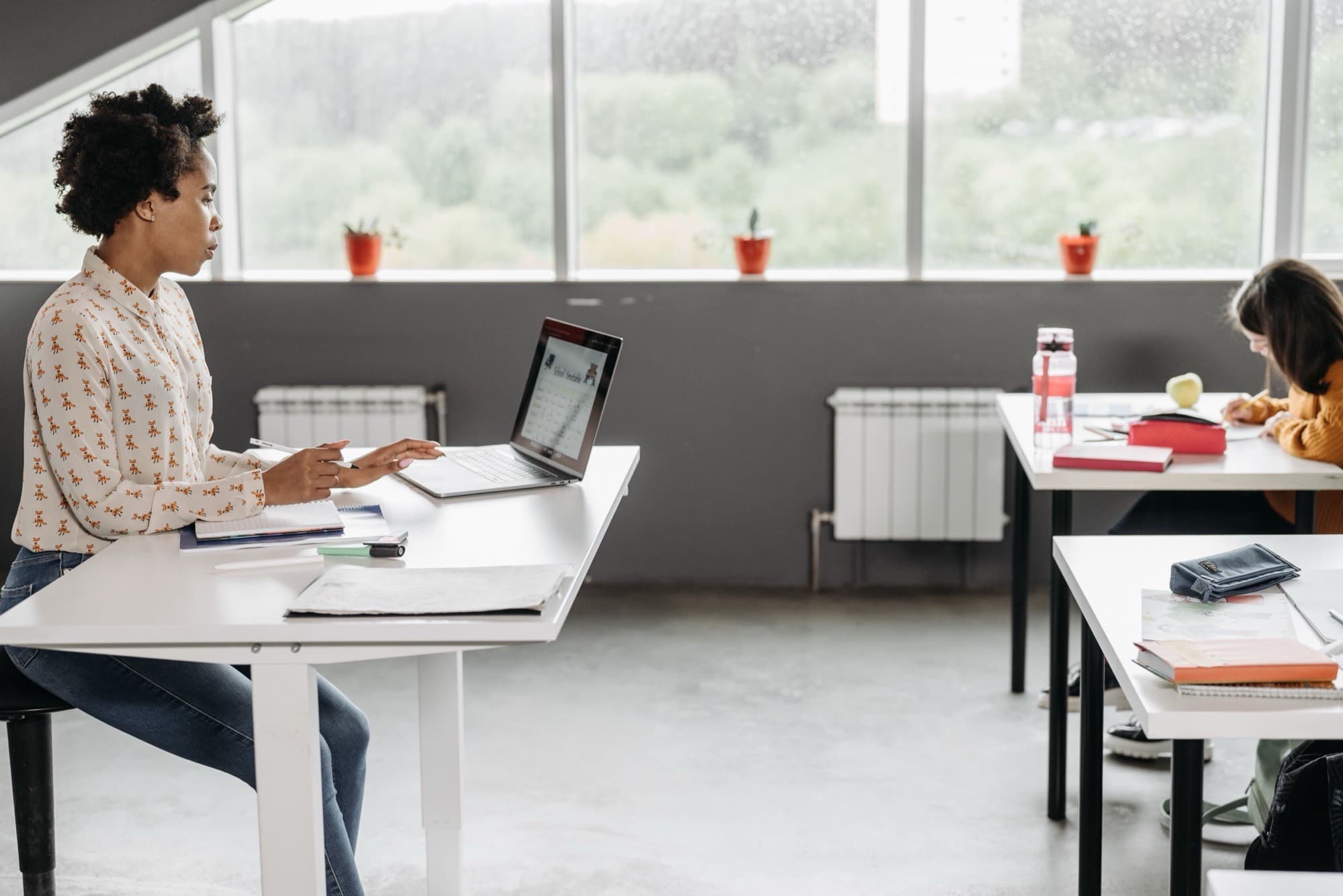 An educator prepares a worksheet on her laptop at a white desk in a bright classroom, ahead of her next lesson.