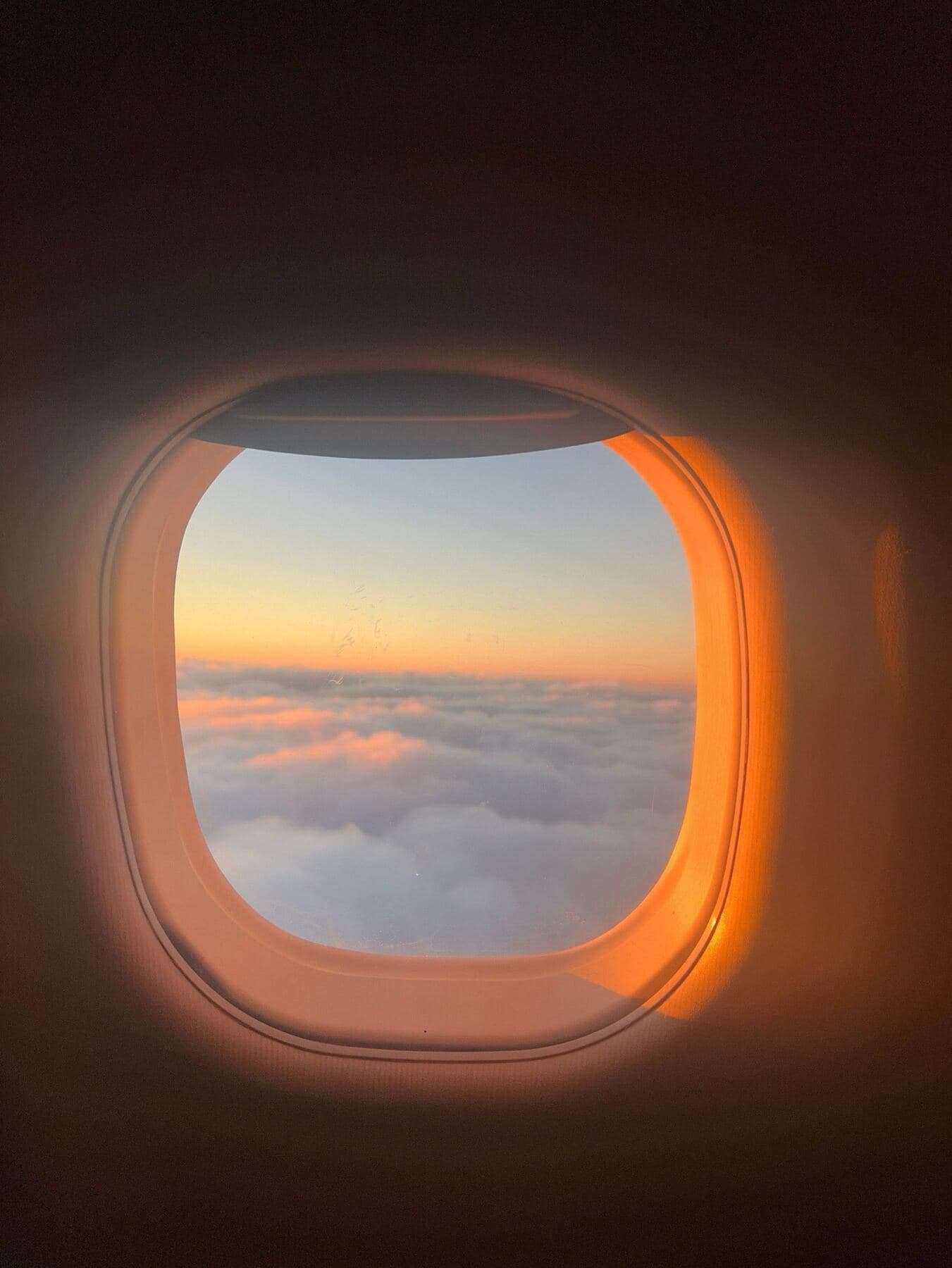 Sunrise over a sea of clouds, seen through a warmly lit aircraft window.