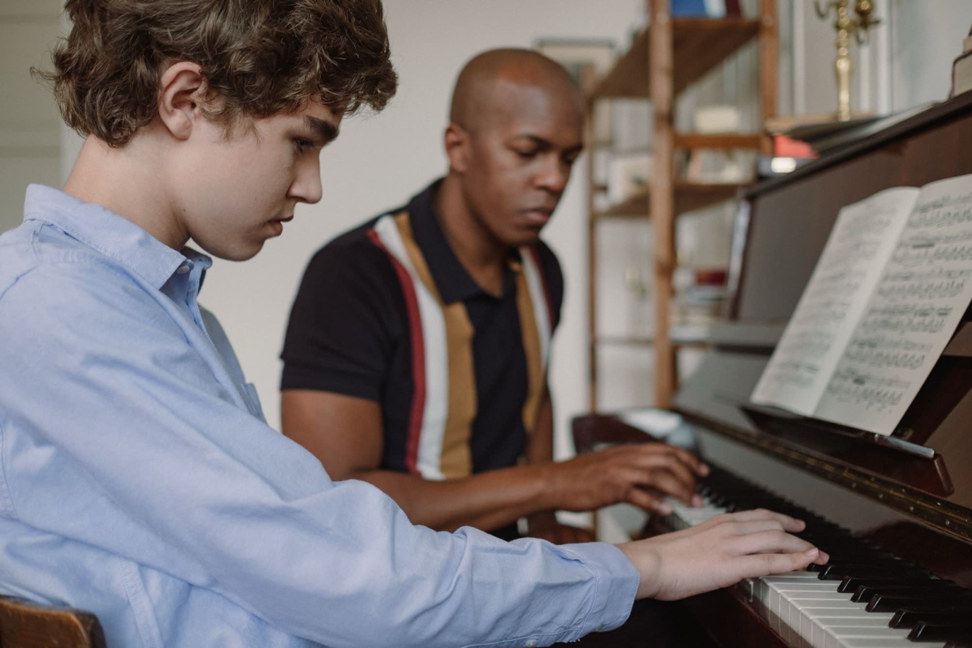 A piano educator plays beside a teenage student, both focused on the keys under an open score.