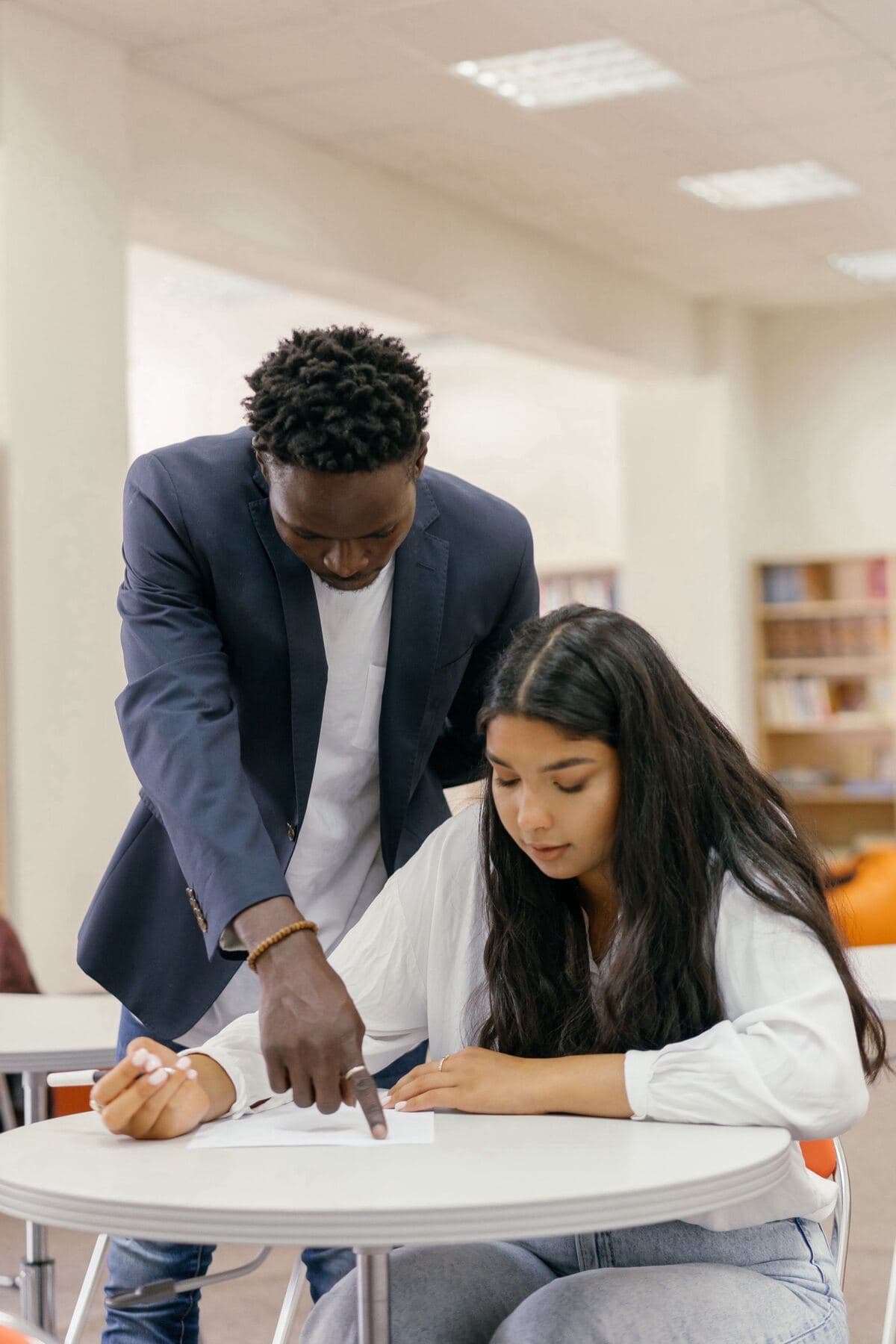 An educator leans over a round table and points to a line on the page while the learner beside him writes her answer.