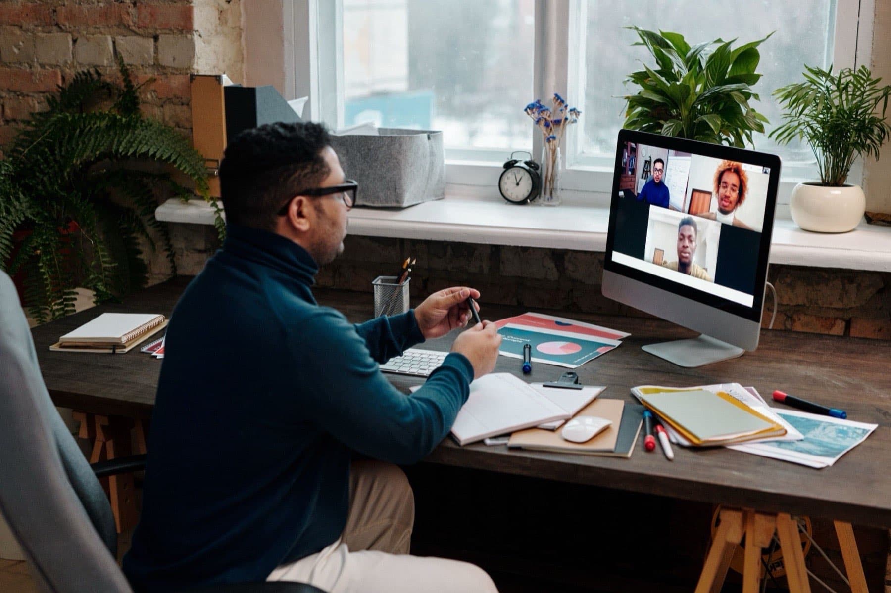 An educator at a desk by a window teaches a partner course live to three learners on screen, notes and markers spread in front of him.