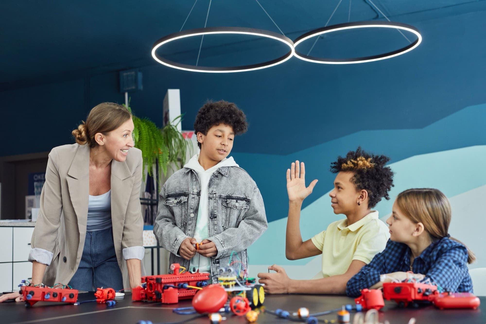 An educator smiles as children build red robot kits at a table, one boy raising his hand to share an idea.
